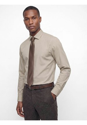Homme portant une chemise beige, une cravate marron, une ceinture marron foncé et un pantalon texturé foncé, debout avec une main dans la poche sur fond blanc.