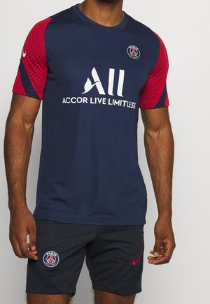 Homme portant un maillot de football bleu marine du Paris Saint-Germain avec des manches rouges et un short assorti avec les logos de l'équipe.