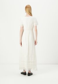 Vestido maxi branco com mangas curtas, cintura elástica e acabamento em renda em camadas na bainha. Tecido texturizado com um design fluido. Calçado preto.