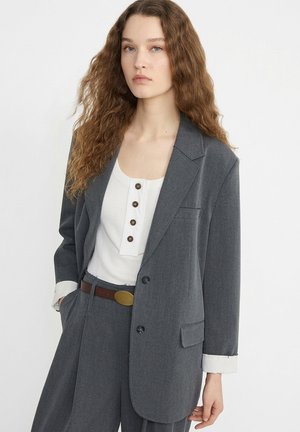 Junge Frau mit langen lockigen Haaren, die eine graue übergroße Anzugjacke und Hose, ein weißes, geknöpftes Oberteil und einen braunen Gürtel mit Messingschnalle trägt.