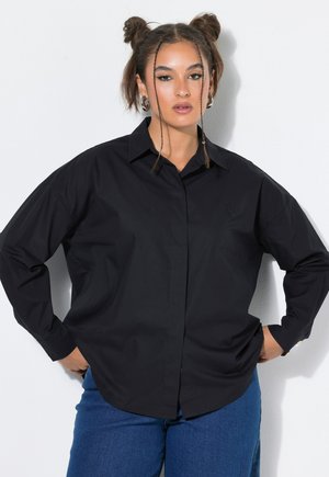 Vrouw met twee gevlochten knotjes die een losse zwarte lange mouwen blouse en blauwe jeans draagt, staand tegen een witte achtergrond.