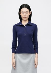 Polo cu mâneci lungi, de culoare bleumarin, cu guler cu nasturi, accesorii argintii și croială ajustată. Asortat cu o fustă gri deschis.