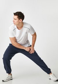 Biała sportowa koszulka z szarymi akcentami, zestawiona z granatowymi spodniami dresowymi; model ma na sobie białe sneakersy; gładkie tekstury, luźny krój.