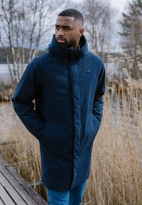 Parka impermeabile blu navy con colletto alto, zip frontale e due tasche laterali. Presenta un logo del brand discreto sul petto e una vestibilità comoda.