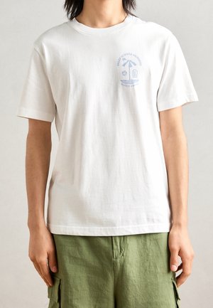 Personne portant un T-shirt blanc avec un petit motif de parasol de plage bleu et un texte sur le côté gauche de la poitrine, associé à un short cargo vert.