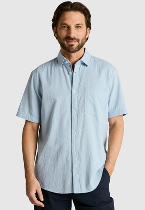 Mann mit kurzen braunen Haaren und Bart, der ein hellblaues kurzärmeliges Hemd mit Brusttasche und dunkle Hosen trägt.