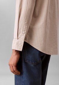 Μανσέτα πουκαμίσου με μακριά μανίκια σε μπεζ και λευκές ρίγες με κουμπί, φορεμένη από άτομο με σκούρα μπλε τζιν, σε απλό γκρι φόντο.