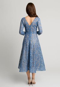 Niebieska koronkowa sukienka z długimi rękawami, dekoltem w kształcie litery V z tyłu i spódnicą do kolan. Koronka ma kwiatowe wzory, a gorset jest dopasowany. Beżowe szpilki.