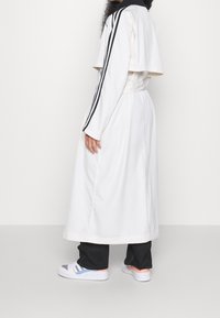 Lång, vit sportrock med korta ärmar, svarta ränder längs axlarna och insvängd midja. Kombinerad med svarta byxor och vita sneakers.