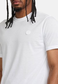 Vit kortärmad t-shirt i mjukt tyg med en cirkulär vit logotyp på bröstet. Slät textur med en klassisk rundhalsad design.