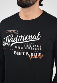 Sort langærmet t-shirt med hvid og orange trykt tekst i forskellige skrifttyper og stilarter, der angiver brandingoplysninger og vintage-elementer.