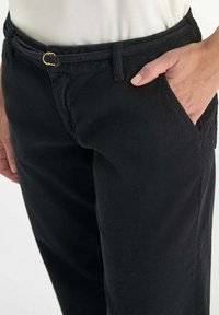 Pantalones de pana negros con una tela texturada, que presentan un cinturón con un lazo de metal dorado y bolsillos laterales, exhibidos a la altura de la cintura.