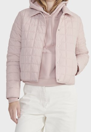 Kvinde iført en lyserød, quiltet jakke over en matchende lyserød hættetrøje og hvide bukser, stående med den ene hånd i lommen.