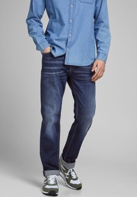 Jeans din denim albastru expuși alături de o cămașă cu butoni de culoare albastru deschis. Jeansii au o croi ajustat și o estompare subtilă; asortați cu adidași verzi și albi.