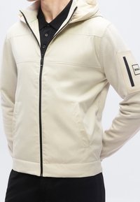 Homme portant une veste à capuche beige zippée par-dessus une chemise noire à col, les mains dans les poches de la veste.