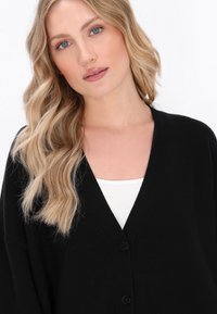 Cardigan negru cu un design în formă de V, având două butoane în față. Materialul pare moale și texturat, purtat peste un top alb.