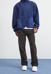 Marinblå fleecetröja med hög krage, dragkedjestängning och tvåfärgade fickor; tillsammans med mörkgrå byxor och vita skor på en blå bakgrund.