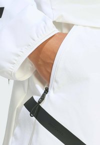 Bijela sportska jakna s džepom na patentni zatvarač, elastičnim manšetama i crnim naglašenim remenom s natpisom "SPORTSWEAR." Mekana, glatka tekstura tkanine.