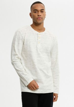Joven con pelo corto que lleva una camiseta henley de manga larga gris claro y pantalones negros, de pie contra un fondo liso.