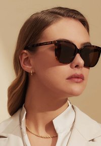 Lunettes de soleil écaille de tortue brun avec des verres foncés et une forme carrée, présentant des détails dorés sur les branches. Blouse de couleur claire en arrière-plan.