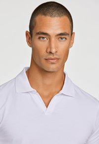 Valkoinen poolopaita, jossa on klassinen kaulus, sileä pinta ja kolmen napin nappilista. Paitaa käyttää lyhyttukkainen malli neutraalilla taustalla.