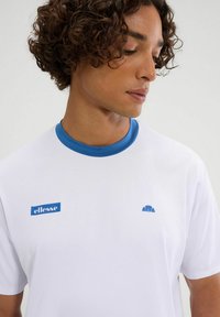 Weißes, kurzärmliges Sporthemd mit blauer Kragen, mit "ellesse"-Logo und kleinen blauen Akzenten am Kragen. Glatte Stofftextur.