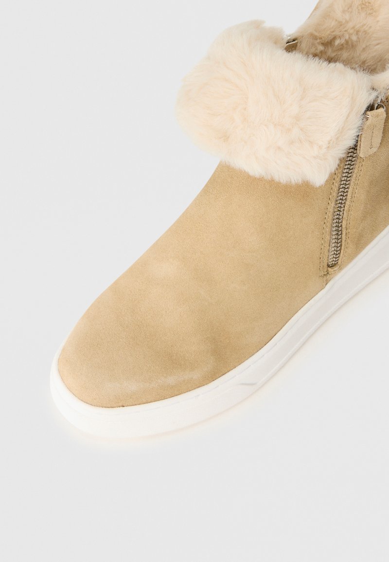 Beige mockask ankle boot med vikbar krämfärgad fuskpäls, sidodragkedja och vit gummisula på en vit bakgrund.