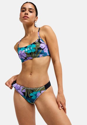 Kobieta w fioletowym i morskim bikini w kwiaty, trzyma lewą ręką bok dołu bikini na jednolitym jasnym tle.