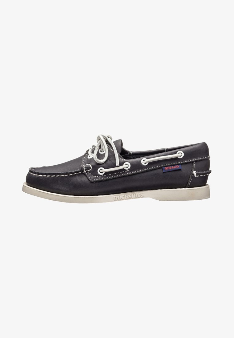Zapato de barco de cuero negro con cordones blancos, suela de goma blanca y costuras visibles. Presenta una pequeña etiqueta roja de Sebago en el lateral.