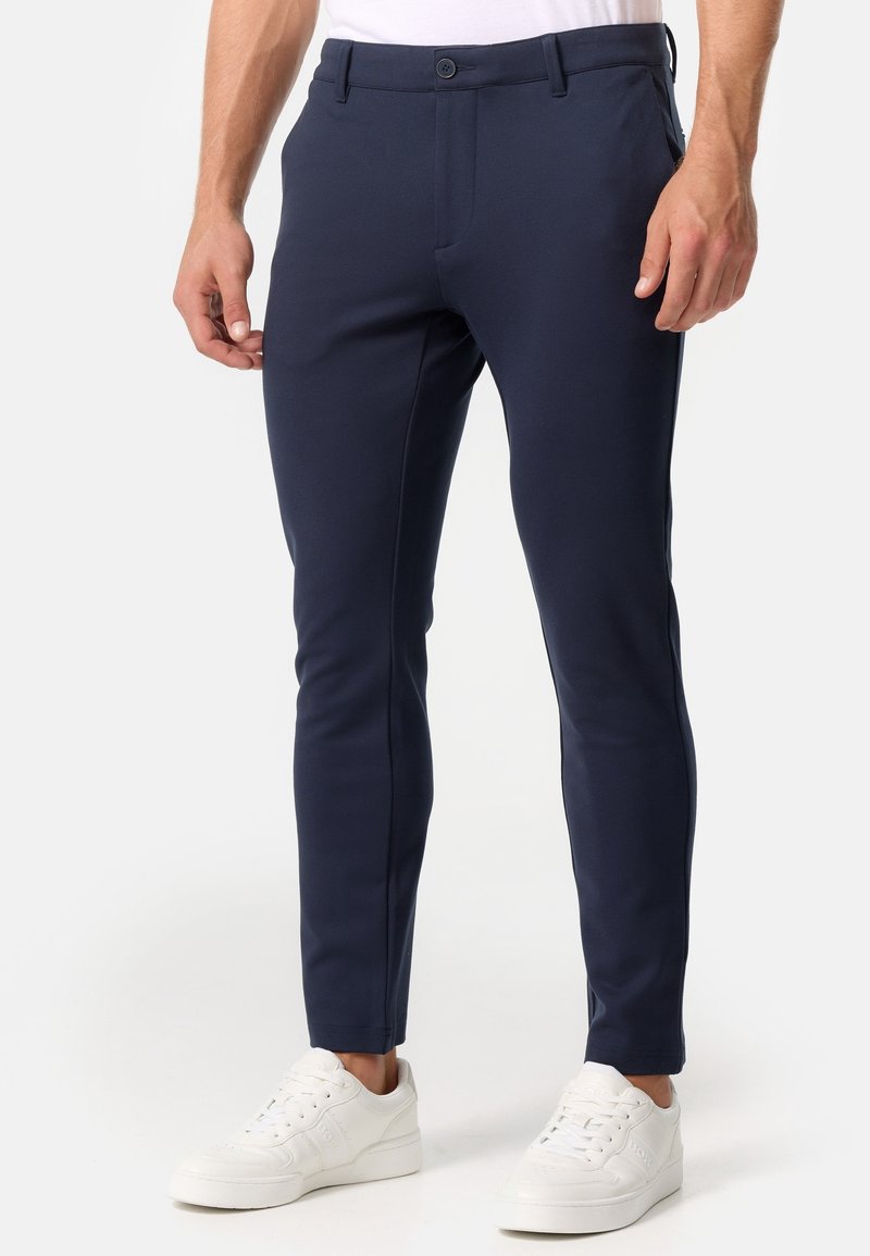 Homme portant un pantalon bleu marine ajusté et des baskets blanches, debout devant un fond clair uni.