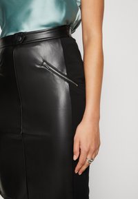 Svart blyertskjol med läderframsida, tygbak, sidoficka med dragkedja och figurnära passform. Sidentopp i ljus teal-färg.