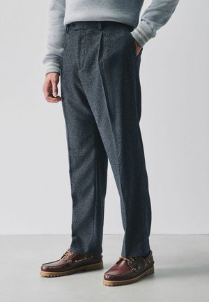 Mann trägt dunkel strukturierte Hose, braune Leder-Bootsschuhe und ein hellgraues Sweatshirt mit gestreiften Bündchen, steht auf einem hellen Boden.