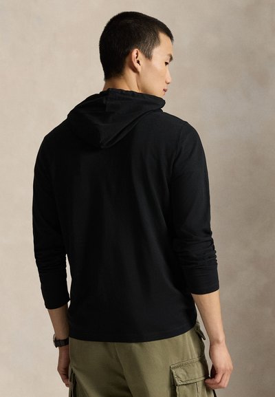 Moški, obrnjen v stran, oblečen v črn dolg rokav s kapuco in olivno zelene hlače cargo na nevtralni podlagi.