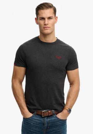 Man draagt een donkergrijs aansluitend T-shirt, een bruine leren riem, een blauwe spijkerbroek en een polshorloge, staat met zijn handen in de zakken.