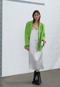 Blazer oversized verde néon sobre um vestido slip de cetim prateado, combinado com botas pretas de salto grosso. Uma mala minimalista prateada complementa o visual.