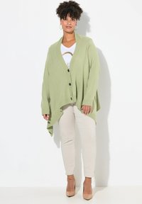 Grön stickad kavaj med V-ringning, långa ärmar och en avslappnad passform. Svarta knappar och en asymmetrisk nederkant. Bärs över en vit topp och beigefärgade byxor.