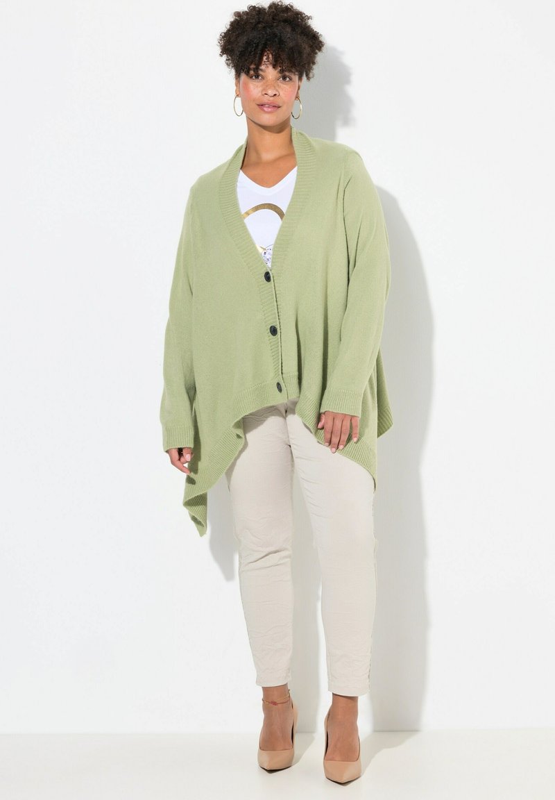 Grön stickad kavaj med V-ringning, långa ärmar och en avslappnad passform. Svarta knappar och en asymmetrisk nederkant. Bärs över en vit topp och beigefärgade byxor.