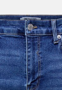 Nærbilde av blå denim jeans med synlig toppknapp, beltehempe, foran lomme-søm og merkelapp delvis synlig inne i livvidden.