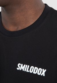 Sort bomulds t-shirt med rund hals, der har et hvidt "SMILODOX" logo på forsiden. Glat tekstur, minimalistisk design.