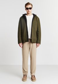 Olivgrüne Zip-Jacke mit schwarzem Innenfutter, kombiniert mit beigen Hosen und braunen Slippern; das Modell trägt schwarze Sonnenbrillen.