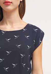 Marinblus med rund halsringning och korta ärmar, prydd med vita kolibri-mönster, tillverkad av lättviktig tyg.