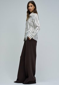 Camisa blanca de manga larga con rayas verticales negras, combinada con pantalones marrones anchos. La camisa tiene detalles de botones y un corte relajado.