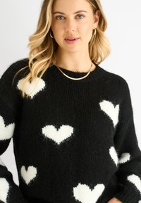Black knitted sweater with white heart patterns; ribbed neckline and cuffs; soft, textured fabric; relaxed fit.