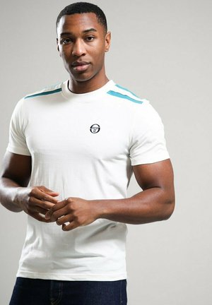 Giovane uomo che indossa una t-shirt bianca con strisce turchesi sulle spalle e un piccolo logo sul petto, in piedi davanti a uno sfondo semplice.