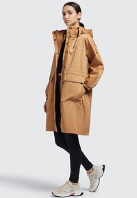 Světle hnědá nepromokavá parka s kapucí, se zapínáním na zip, dvěma velkými předními kapsami a elastickými manžetami. Nositá přes černé legíny a boty.