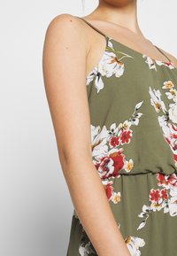 Kukkajumpsuit oliivinvihreästä kankaasta, jossa on valkoisia ja punaisia kukkia. Ohut spagettiolkaimet ja kapea vyötärödesign. Sileä pinta.