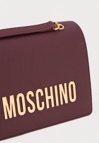 Burgundia faux nahast käsitasku kuldse "MOSCHINO" kirjaga ja struktureeritud disainiga. Omab õhukest käepidet ja metallist aksessuaare.