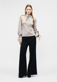 Femme portant un haut gris sans manches avec des découpes aux épaules et des manches nouées, associé à un pantalon large noir et des chaussures noires, debout avec une main dans la poche.