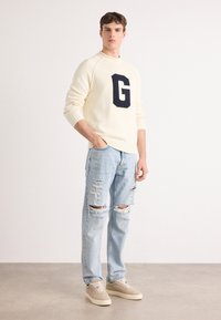Cream sweater with a large navy "G" logo, paired with light blue ripped jeans and beige sneakers. Relaxed fit with ribbed cuffs.