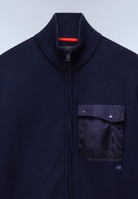 Marinblå stickad zip-jacka med hög krage och strukturerad tyg. Inkluderar en mörk nylonframlapp med snap-stängning.